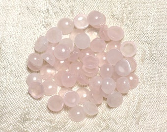 May include: A pile of pale pink rose quartz cabochons, smooth and polished, with a slightly translucent appearance. The stones are arranged in a random pattern on a white fabric background.