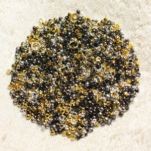 May include: A close-up of a pile of small, round beads in gold, silver, and black colors. The beads are arranged in a circular shape on a white fabric background.