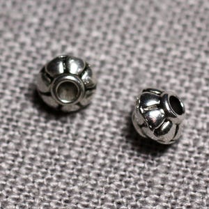 Peut inclure: Deux perles en métal argenté avec un motif décoratif en forme de fleur. Les perles ont un trou au centre pour être enfilées.