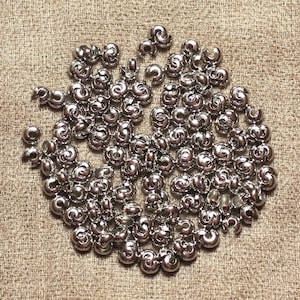 May include: A collection of shiny, silver-colored metal beads with a spiral design. The beads are scattered on a textured, tan fabric background. These decorative beads are suitable for jewelry making and craft projects.