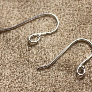 Peut inclure: Deux crochets d'oreille en argent, chacun avec une petite boucle en haut et une extrémité arrondie. Les crochets sont posés sur une surface en toile de jute brune.