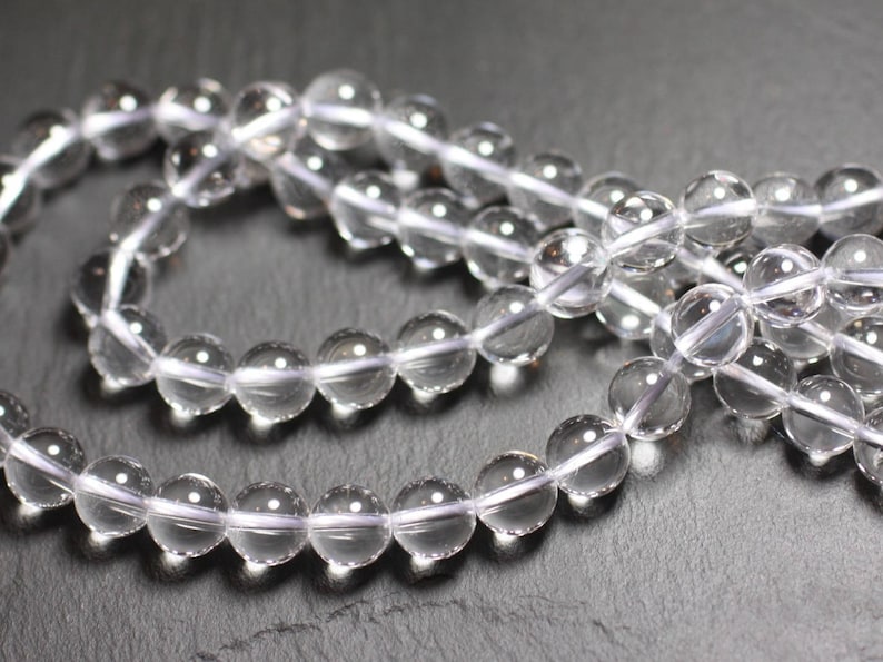 Puede incluir: Primer plano de cuentas de cristal de cuarzo transparente. Las cuentas son esf&eacute;ricas y transparentes, ensartadas en un cord&oacute;n blanco. Cada cuenta mide unos 8 mm de di&aacute;metro. El fondo es una superficie oscura y texturizada.