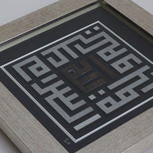 May include: A framed artwork featuring a silver and black geometric design with Arabic calligraphy. The artwork is framed in a light wood frame.