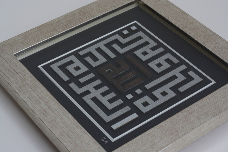 May include: A framed artwork featuring a silver and black geometric design with Arabic calligraphy. The artwork is framed in a light wood frame.