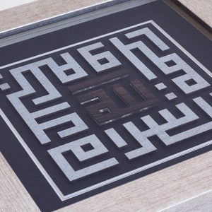 May include: A framed piece of Islamic calligraphy art with a silver and black color scheme. The calligraphy is in a square frame with a black background and a silver border. The calligraphy is in Arabic script and is surrounded by a silver border.