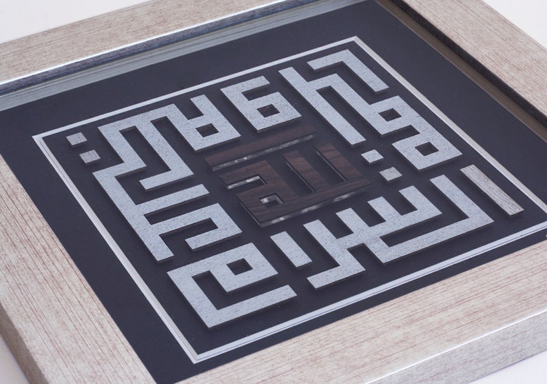 May include: A framed piece of Islamic calligraphy art with a silver and black color scheme. The calligraphy is in a square frame with a black background and a silver border. The calligraphy is in Arabic script and is surrounded by a silver border.