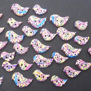 May include: A collection of colorful bird-shaped buttons. Each button features a unique pattern of purple, blue, yellow, and pink, with two small holes for sewing. The buttons are made of a smooth, glossy material.