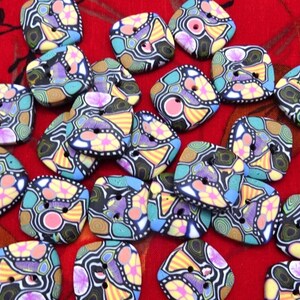 May include: A selection of square, decorative buttons with a vibrant, abstract pattern. The buttons display a variety of colours such as yellow, purple, blue, and black, with two or four holes for sewing. The buttons are placed on a red surface.