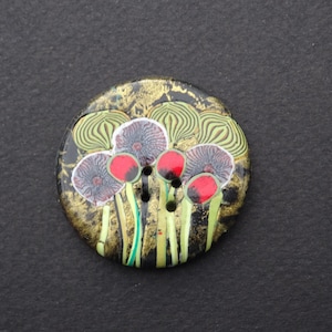 Puede incluir: Un botón redondo con un diseño floral. El botón presenta un fondo negro con detalles dorados. Las flores tienen pétalos rojos y morados con tallos y hojas verdes. El botón tiene tres agujeros para coser.