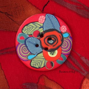 May include: A round, flat button with a vibrant red base featuring a multi-colored floral and leaf pattern. The design includes blue-striped leaves, purple swirls, and a central red flower motif with two visible holes for attachment.
