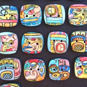 Peut inclure: Une collection de boutons carrés présentant des motifs complexes peints à la main rappelant des glyphes anciens. Chaque bouton possède deux trous centraux et affiche une combinaison unique de teintes jaunes, bleues, rouges et orange.