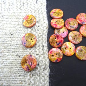 Può includere: Una collezione di bottoni rotondi e colorati, ciascuno con due fori. I bottoni hanno un effetto marmorizzato con sfumature di giallo, rosa e viola. Alcuni bottoni sono posizionati su un tessuto a maglia grigio chiaro, altri su una superficie nera.