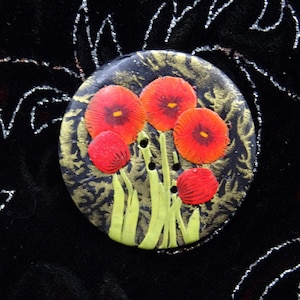 Peut inclure: Un bouton rond noir avec un motif floral peint. Le bouton présente cinq coquelicots rouges avec des centres jaunes et des tiges vertes sur un fond doré et noir. Le bouton a trois trous pour la couture.