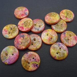 Puede incluir: Una colección de botones redondos de dos agujeros con una superficie moteada y multicolor que presenta salpicaduras vibrantes de rosa, amarillo y naranja. Cada botón tiene un acabado brillante y texturizado con patrones orgánicos y arremolinados.
