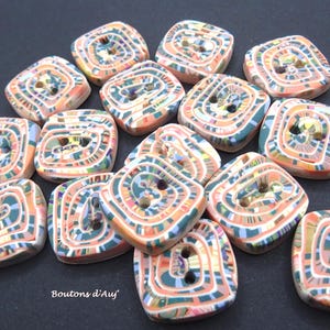 Puede incluir: Una colección de botones cuadrados con un patrón de remolinos en tonos melocotón, verde azulado, blanco y gris oscuro. Cada botón tiene dos agujeros centrales para su fijación. El texto "Boutons d'Auj" está impreso en blanco en la esquina inferior izquierda.