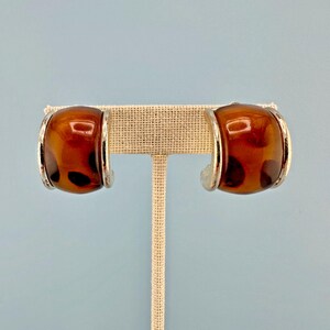 May include: A pair of half-hoop earrings featuring a smooth, polished amber-colored tortoiseshell resin inlay set within a shiny silver-toned metal frame. The earrings have a curved, chunky design with a glossy finish and dark brown spots.