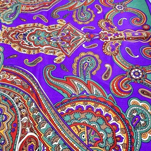 May include: A fabric featuring a vibrant purple background with a detailed, multi-colored paisley pattern. The design includes intricate swirls, teardrop shapes, and floral motifs in shades of orange, teal, yellow, red, and white throughout.