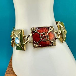 May include: A gold-tone link bracelet featuring a central square plaque with red enamel and a floral motif, flanked by rectangular green enamel links with blue and green accents. The design includes small clear crystals set into the metal frame.