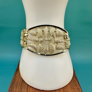May include: A gold-tone metal cuff bracelet featuring a wide, oval-shaped center panel with a textured, leaf-like pattern. The bracelet is constructed with hinged side links that connect to the central decorative piece, creating a structured design.