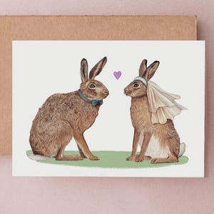 May include: A greeting card featuring two brown hares. One hare wears a blue bow tie, the other a veil. A purple heart floats between them. The card is on a brown envelope, set against a pink background.