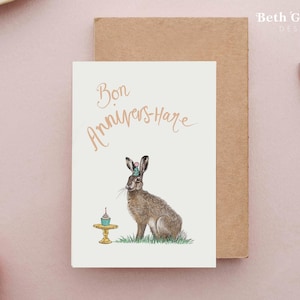 May include: A white card with the text "Bon Annivers-Hare" in brown script. A brown hare with a blue and pink party hat is sitting on a green patch of grass next to a small yellow cake stand with a cupcake on it.