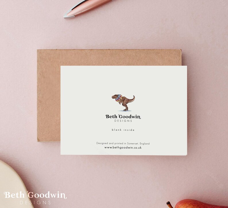 May include: A white card with a cartoon illustration of a Tyrannosaurus Rex wearing a colorful flower garland. The text "Beth Goodwin, DESIGNS" is below the illustration. The text "blank inside" is below the logo. The text "Designed and printed in Somerset, England" is below the text "blank inside". The text "www.bethgoodwin.co.uk" is below the text "Designed and printed in Somerset, England".