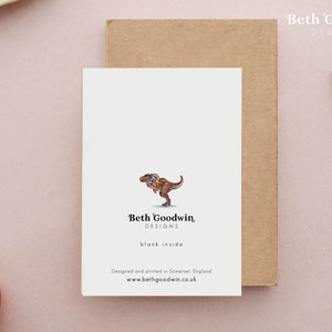K&ouml;nnte beinhalten: Eine Gru&szlig;karte mit einer Cartoon-Illustration eines Tyrannosaurus Rex, der eine bunte Blumenkette tr&auml;gt. Die Karte ist innen leer und hat den Text "Beth Goodwin, DESIGNS" und "blank inside" auf der Vorderseite gedruckt. Die Karte wurde in Somerset, England, entworfen und gedruckt. Die Website-Adresse "www.bethgoodwin.co.uk" ist ebenfalls auf der Vorderseite gedruckt.