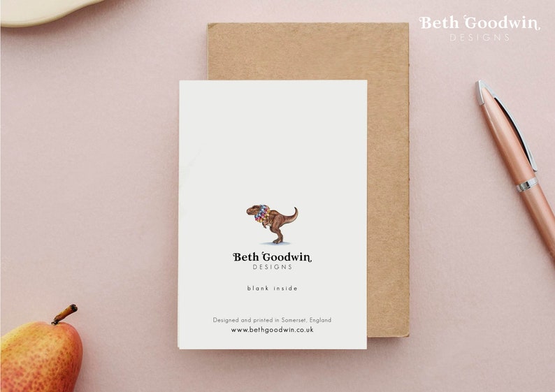 K&ouml;nnte beinhalten: Eine Gru&szlig;karte mit einer Cartoon-Illustration eines Tyrannosaurus Rex, der eine bunte Blumenkette tr&auml;gt. Die Karte ist innen leer und hat den Text "Beth Goodwin, DESIGNS" und "blank inside" auf der Vorderseite gedruckt. Die Karte wurde in Somerset, England, entworfen und gedruckt. Die Website-Adresse "www.bethgoodwin.co.uk" ist ebenfalls auf der Vorderseite gedruckt.