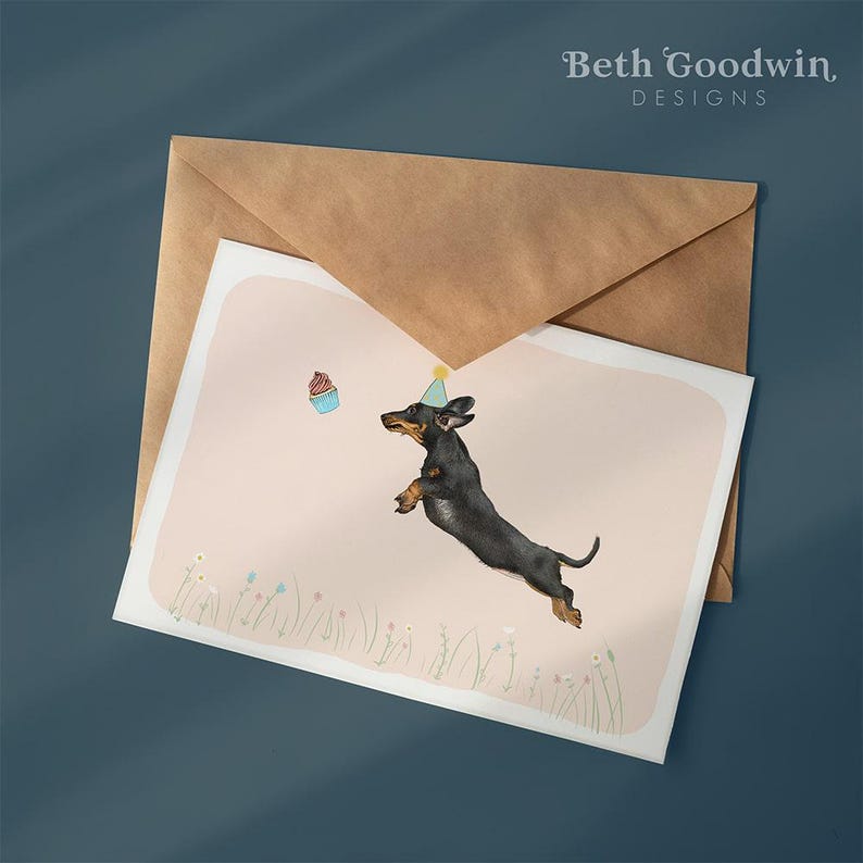 May include: Greeting card featuring a watercolor illustration of a dachshund wearing a party hat, jumping towards a cupcake. The card is on a pink background with floral accents, inside a brown envelope. The text "Beth Goodwin Designs" is at the top.
