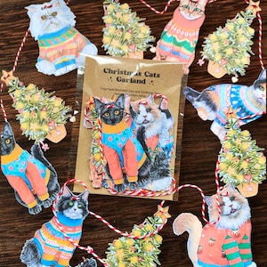 May include: A festive Christmas Cats Garland featuring illustrated cats in sweaters and Christmas trees. The cats are in various colours and patterns, strung together with red and white twine. The packaging reads "Christmas Cats Garland."