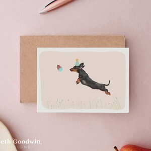 May include: Greeting card featuring a black and brown dachshund wearing a party hat, leaping towards a cupcake. The illustration is set against a light pink background with a brown envelope. The card has the text "Beth Goodwin DESIGNS" at the bottom.