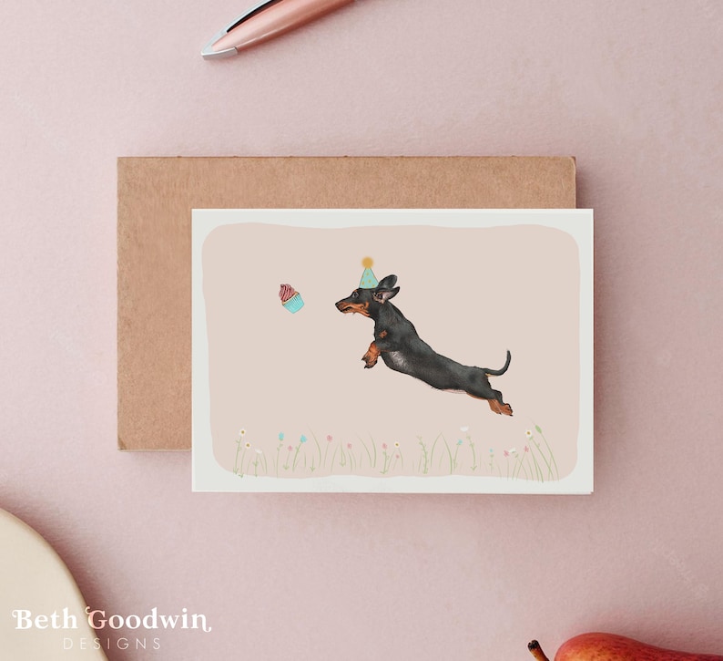 May include: Greeting card featuring a black and brown dachshund wearing a party hat, leaping towards a cupcake. The illustration is set against a light pink background with a brown envelope. The card has the text "Beth Goodwin DESIGNS" at the bottom.