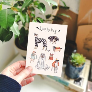 Op de afbeelding: Een kaart met de tekst "Spooky Dogs" en illustraties van honden in Halloween-kostuums. De honden zijn afgebeeld als zebra, spin, spook en andere griezelige personages. De kaart wordt door een persoon vastgehouden.