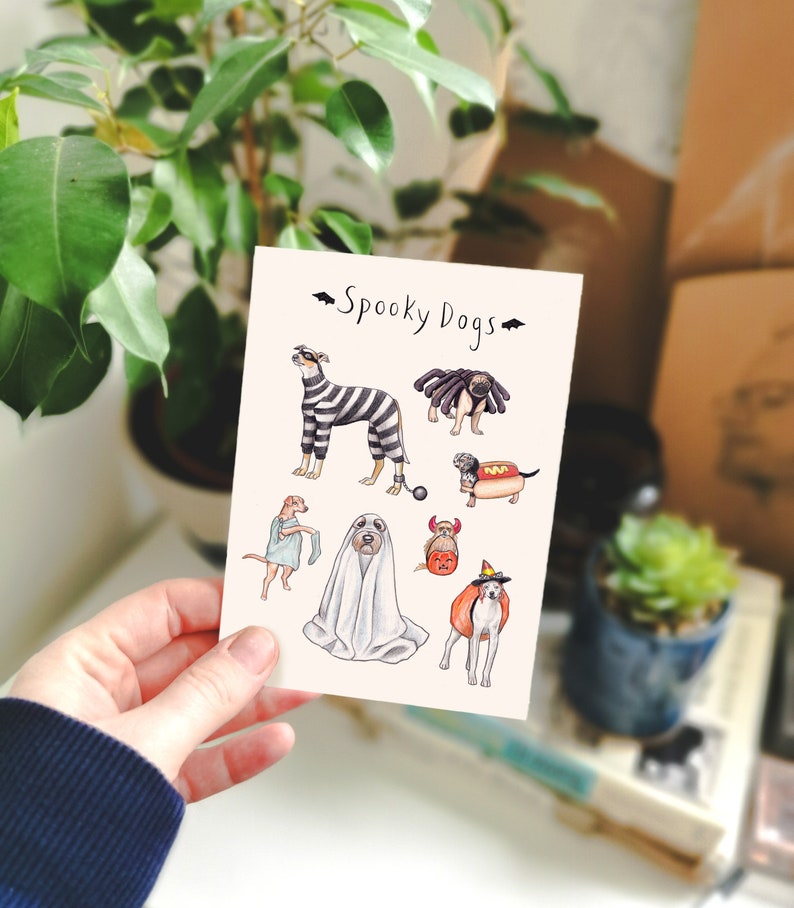 Op de afbeelding: Een kaart met de tekst "Spooky Dogs" en illustraties van honden in Halloween-kostuums. De honden zijn afgebeeld als zebra, spin, spook en andere griezelige personages. De kaart wordt door een persoon vastgehouden.