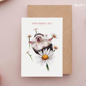 K&ouml;nnte beinhalten: Eine wei&szlig;e Gru&szlig;karte mit einem fr&ouml;hlichen Igel, der eine G&auml;nsebl&uuml;mchen festh&auml;lt. Die Karte lautet "Happy Mother's Day" in rosa Schrift.