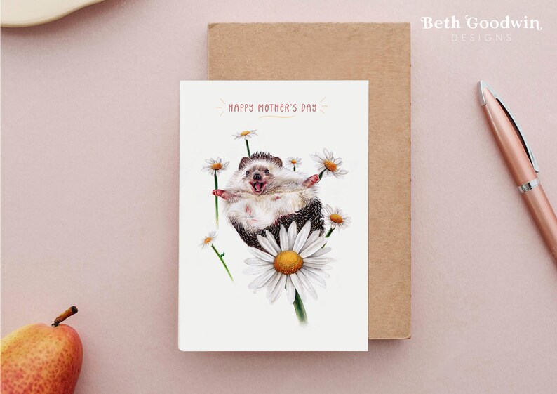 K&ouml;nnte beinhalten: Eine wei&szlig;e Gru&szlig;karte mit einem fr&ouml;hlichen Igel, der eine G&auml;nsebl&uuml;mchen festh&auml;lt. Die Karte lautet "Happy Mother's Day" in rosa Schrift.
