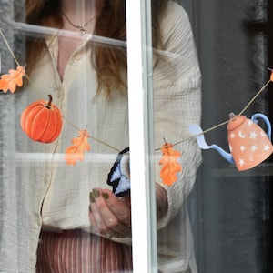Puede incluir: Una guirnalda decorativa con recortes de papel de un búho blanco, calabazas naranjas, hojas de roble naranjas, un conejo marrón y una tetera azul claro con patrones de estrellas y lunas blancas, todo ensartado en un cordel fino de color claro.