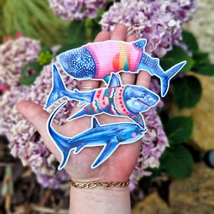 May include: Three shark stickers are held in a hand. The top shark is a whale shark wearing a pink sweater with rainbow stripes. The middle shark has a blue body and wears a sweater with a colorful pattern. The bottom shark is blue.