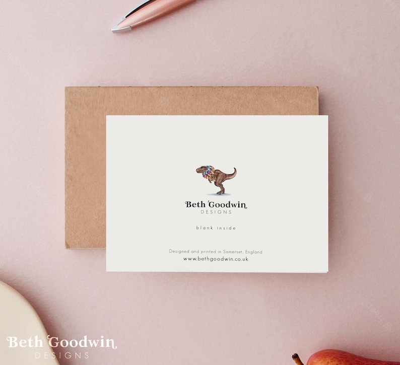 May include: A greeting card with a watercolor illustration of a dinosaur wearing a floral collar. The card is blank inside and features the text "Beth Goodwin DESIGNS" and website information. The card is on a light pink surface.