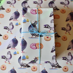May include: A rectangular gift wrapped in light grey paper featuring a repeating pattern of pigeons wearing party hats, pink and orange doughnuts, and heart-centred biscuits. The package is tied with a thin, bright blue twine string.