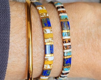 May include: A set of three bracelets consisting of one smooth gold-tone metal bangle and two beaded bracelets. The beaded bracelets feature small rectangular tiles in alternating colors of royal blue, white, mustard yellow, and turquoise with gold spacers.