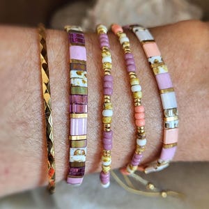 Könnte beinhalten: Ein Set aus fünf Armbändern, bestehend aus einem dünnen, goldfarbenen Metallreif mit strukturierter Oberfläche und vier Perlenarmbändern. Die Perlenmuster kombinieren quadratische und runde Perlen in Rosa, Weiß, Gold und gesprenkeltem Braun.