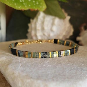 Peut inclure: Bracelet à maillons dorés orné de petites perles rectangulaires dorées, noires et turquoise. Le bracelet est présenté sur une surface blanche. Les perles mesurent environ 0,5 cm de long.