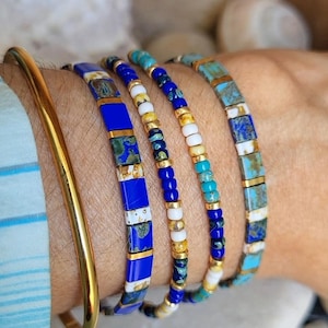 May include: A set of five bracelets including one smooth gold-tone bangle and four beaded bracelets. The beaded designs feature a mix of square and round beads in shades of royal blue, turquoise, white, and gold-tone accents with marbled patterns.