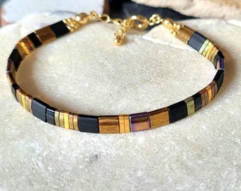 May include: A bracelet featuring a repeating pattern of square-shaped beads in matte black, metallic gold, and iridescent bronze tones. The bracelet includes a gold-toned metal lobster claw clasp and a short extender chain with small gold beads.