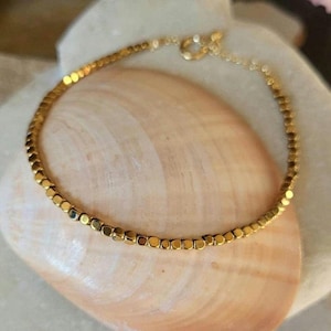 May include: A gold bracelet made of small square beads. The bracelet is on a light brown background.