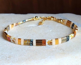 May include: A delicate bracelet composed of alternating rectangular beads in cream, metallic gold, and patterned brown tones. The bracelet features a gold-toned metal lobster clasp and a short extender chain with a small round charm at the end.