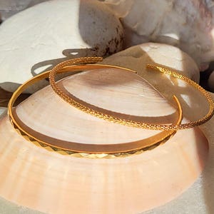 Peut inclure: Deux bracelets en or, l'un avec une surface texturée et l'autre avec un motif en forme de diamant, sont présentés sur une coquille de mer blanche.