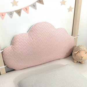 Può includere: Un cuscino rosa a forma di nuvola con un motivo a zig-zag bianco. Il cuscino è in una culla con una piccola pecora di peluche. Uno striscione scandisce il nome "LOUISE" con decorazioni a forma di stella.