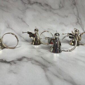 May include: Silver angel napkin rings. Each angel figure is playing a trumpet and has a halo. The napkin rings are made of metal and have a circular design. The angels are standing on a white and gray marble surface.